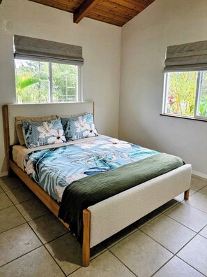 Ein Schlafzimmer mit einem Bett, zwei Fenstern mit Jalousien, einer Holzbalkendecke und einem Fliesenboden.