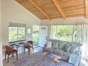 A living room with a wooden ceiling, a sofa, a coffee table, a dining table with chairs, and large windows with curtains. - Koa + Paka: Embrace the elements in style (Keaau)