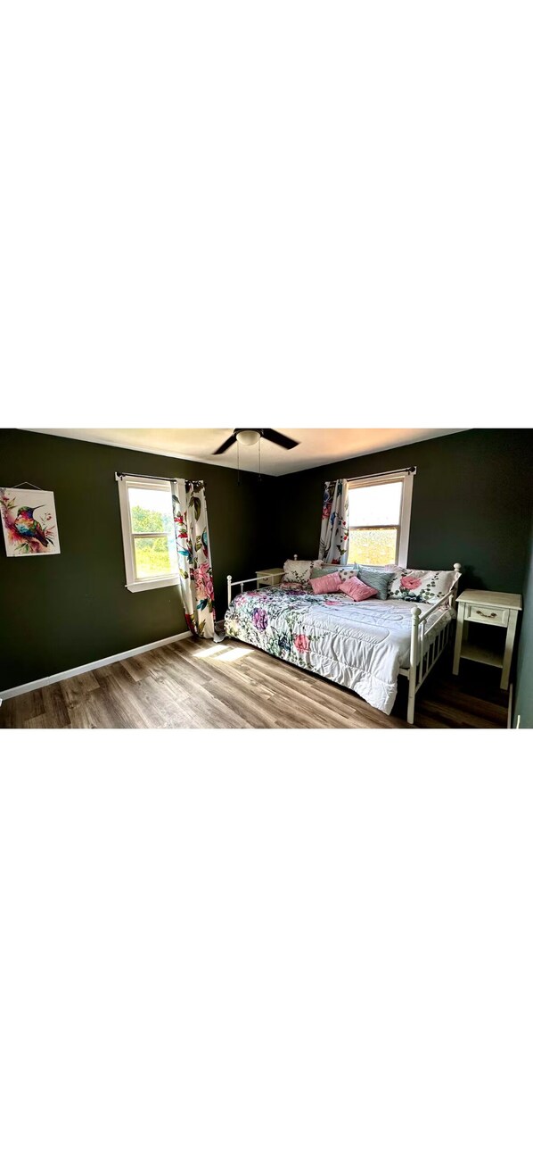 Une chambre à coucher avec un lit, une table de chevet, un ventilateur de plafond et une fenêtre avec des rideaux.
