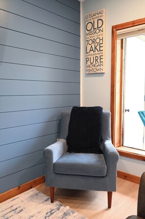 Una habitación en esquina con un sillón gris, suelo de madera, ventana con vistas y una pared con un letrero vertical.