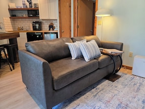 A living room with a sofa, a coffee table, a lamp, and a kitchen area in the background.