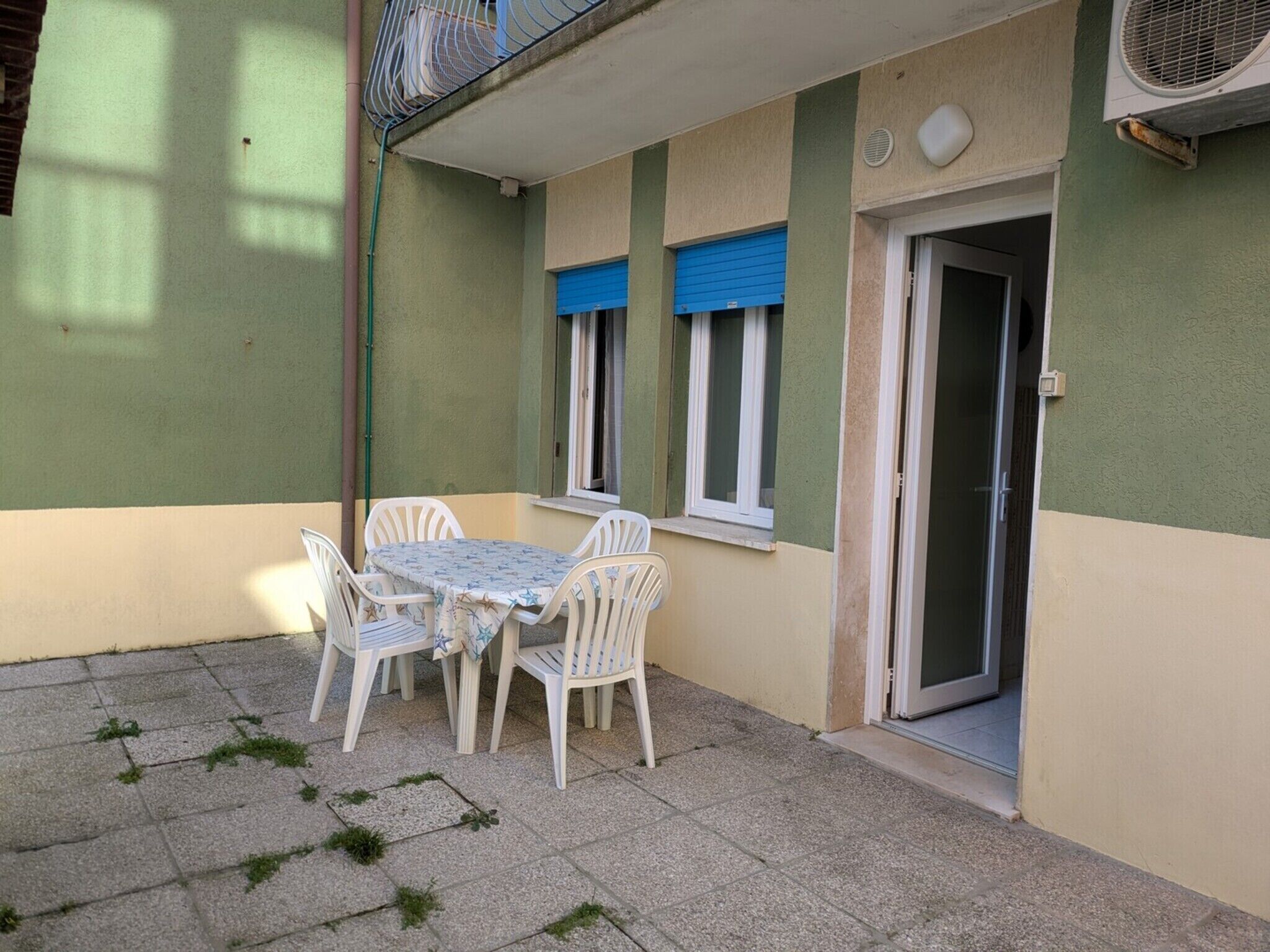 Zona exterior petita amb terrassa, taula i cadires, una finestra amb persianes blaves i una porta corredissa de vidre.