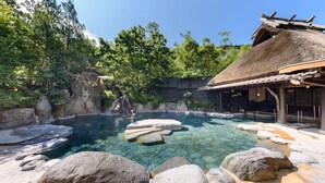 Ein klares Schwimmbecken, umgeben von Felsen und Bäumen, ein traditionelles Gebäude mit einem Strohdach und ein Holzzaun.