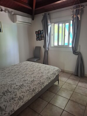 A bedroom with a bed, a chair, a window with curtains, and a wall-mounted air conditioner. - Villa Bleu 2 bedrooms with pool (Sainte-Anne)
