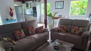 Sala de estar com dois sofás, uma mesa de centro de vidro e vista para uma cozinha e área verde externa.