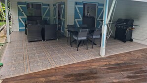 Uma área de varanda coberta com mesa e cadeiras, churrasqueira e fachada azul e branca.