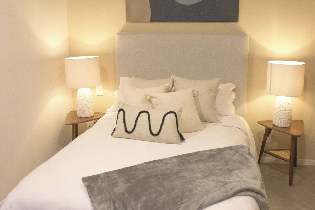 Un lit bien fait, avec une tête de lit, deux tables de chevet avec des lampes et une œuvre d’art accrochée au mur au-dessus du lit.