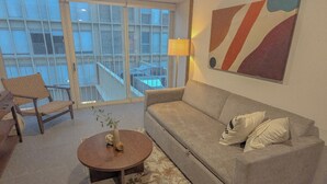 Una sala de estar con un sofá, una mesa redonda de centro, una silla y un ventanal con vistas a edificios.