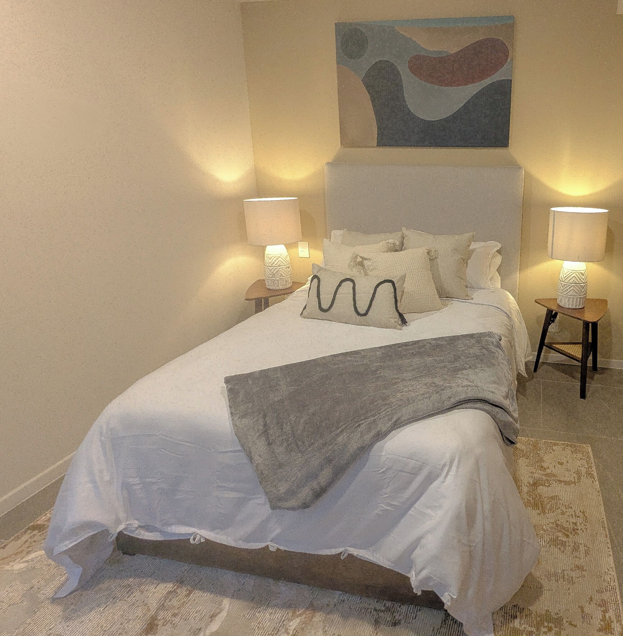 A neatly made bed with pillows and a blanket, two bedside lamps, a rug, and a painting on the wall.