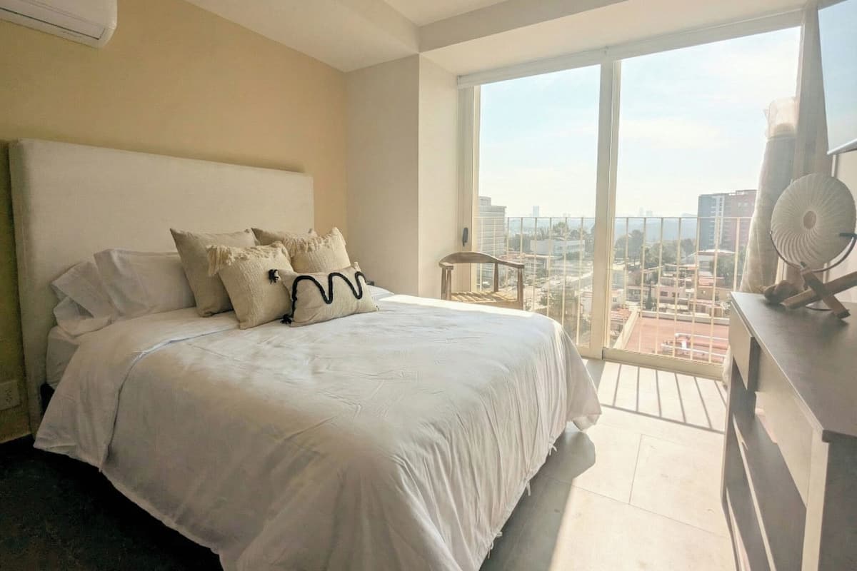 Une chambre avec un grand lit, une vue sur la ville et un balcon équipé d’un ventilateur.