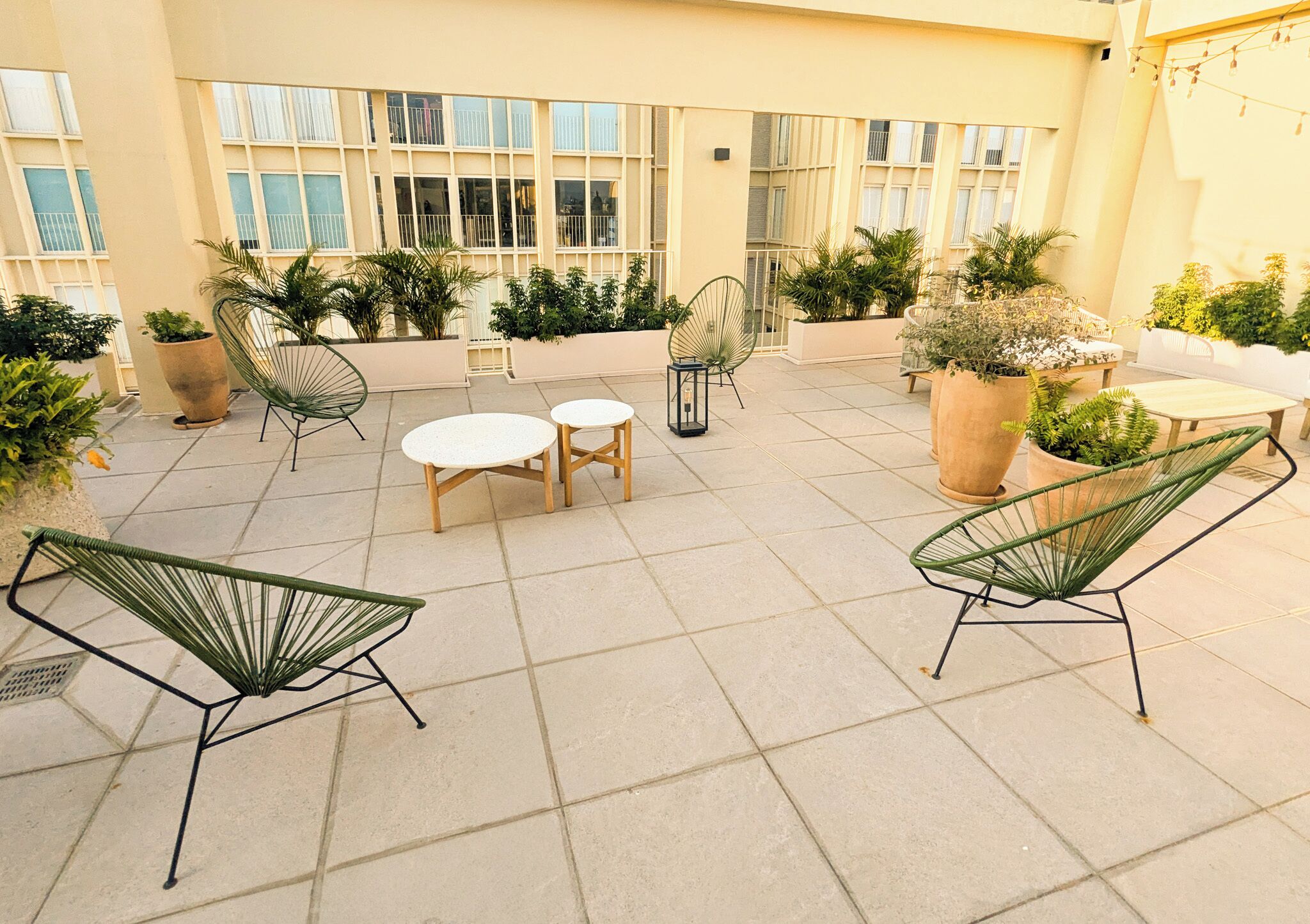 A rooftop patio with outdoor seating, potted plants, and a building with large windows in the background.