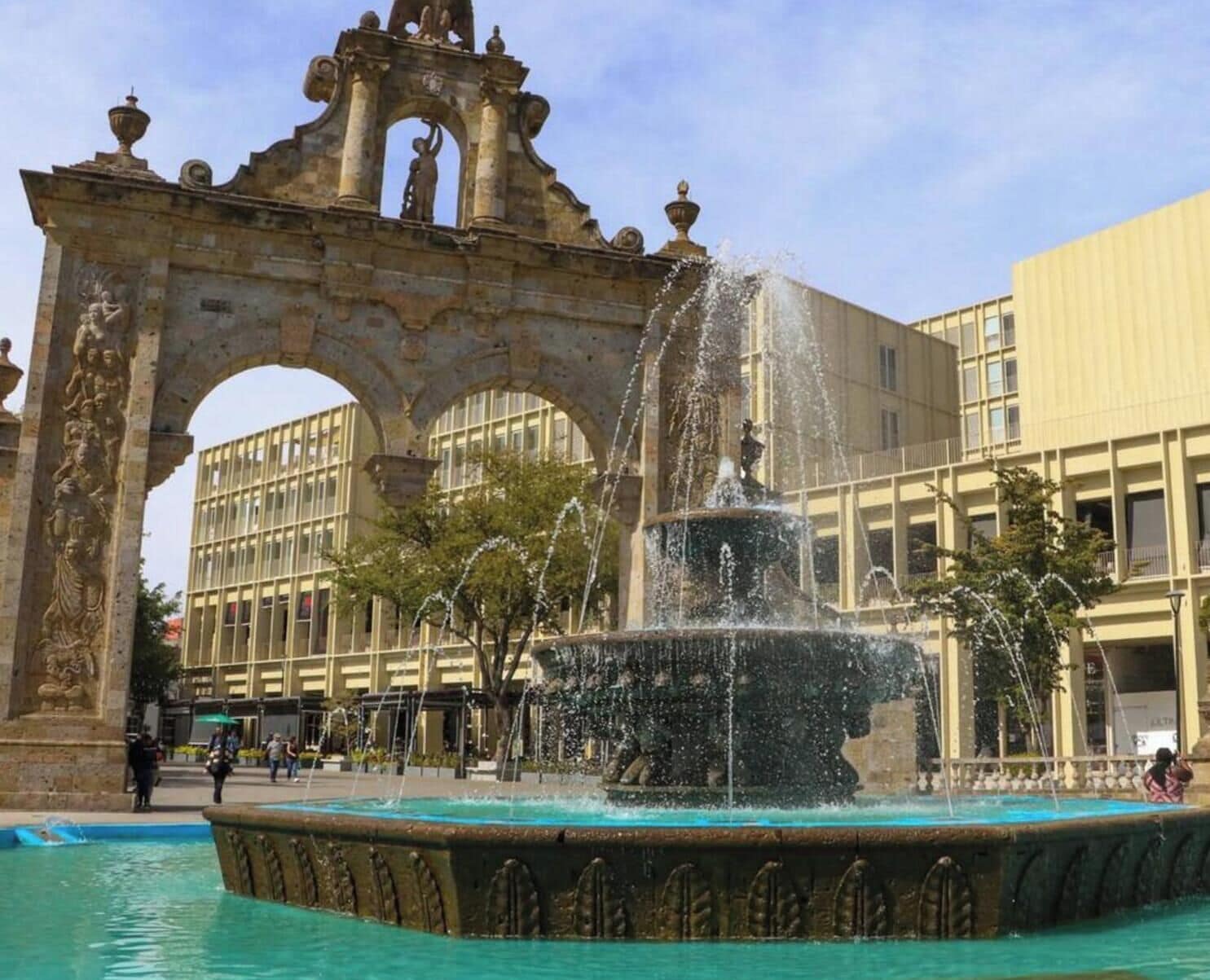 A historic fountain with water jets, an ornate archway, and a modern building in the background.