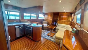 A compact kitchen with a dishwasher, oven, and a small island with a sink. There is a seating area with a cushioned sofa and a small table. - Reel Excuse - Yacht Living (Fort Myers Beach)