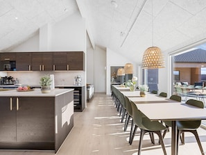 Une cuisine moderne avec des meubles de couleur bois foncé, un îlot central et un coin repas agrémenté de chaises vertes et de luminaires suspendus.
