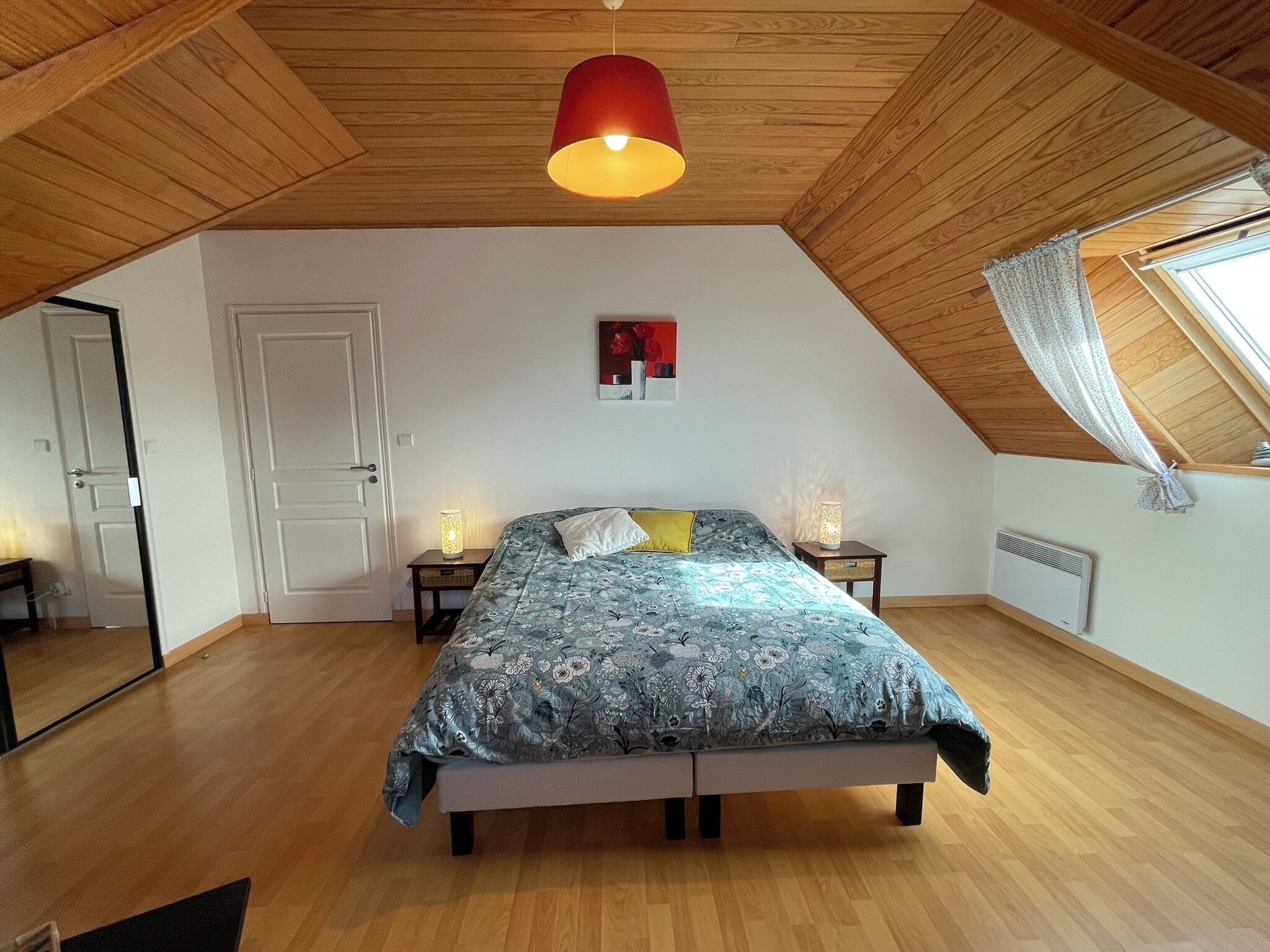 Et soveværelse med skråvæg, en seng med blomstret sengetøj, to sengeborde med lamper, en radiator og et vindue med gardin.
