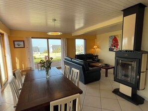 A dining area with a wooden table and chairs, a sofa, a fireplace, and a view of the outdoors. - D6054-Holiday home in Plomodiern with quiet garden and sea view! (Plomodiern)