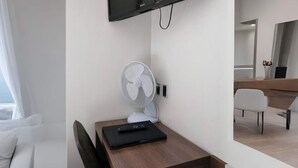 Habitación de hotel con televisión de pantalla plana en la pared, ventilador de pie, mesa de madera con un control remoto y una silla blanca.