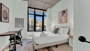 A modern hotel room with a bed, desk, chair, and large windows offering a city view. - Sleek NoLibs 1BR Close to Hotspots | Sleeps 4 (Philadelphia)