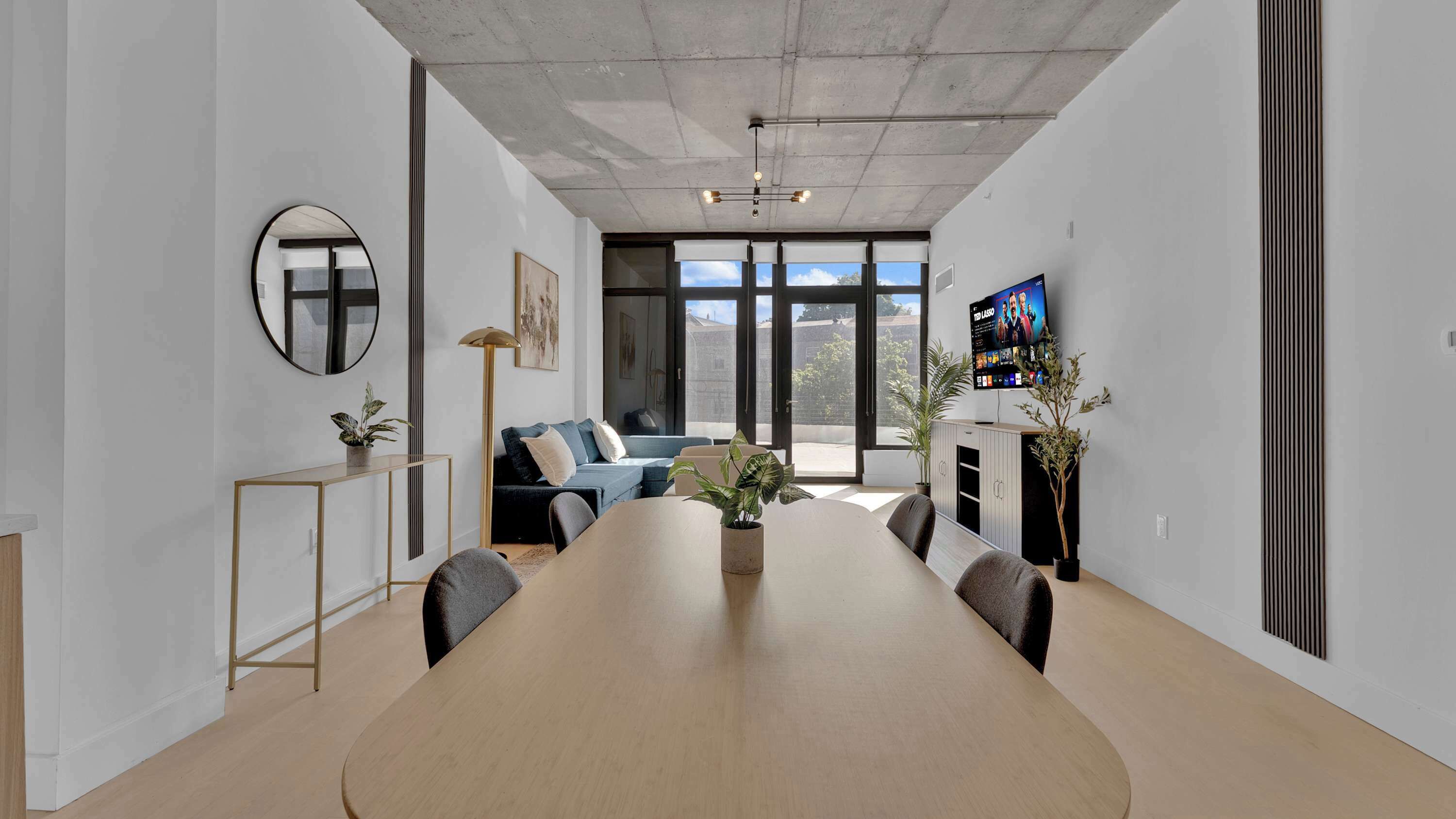 Une salle de réunion moderne, dotée d’une longue table, de chaises, d’un grand miroir rond, d’un canapé et d’un téléviseur fixé au mur.