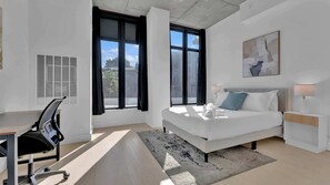 A modern hotel room with a bed, desk, chair, and large windows. - Trendy NoLibs 1BR Near City Spots | Sleeps 4 (Philadelphia)