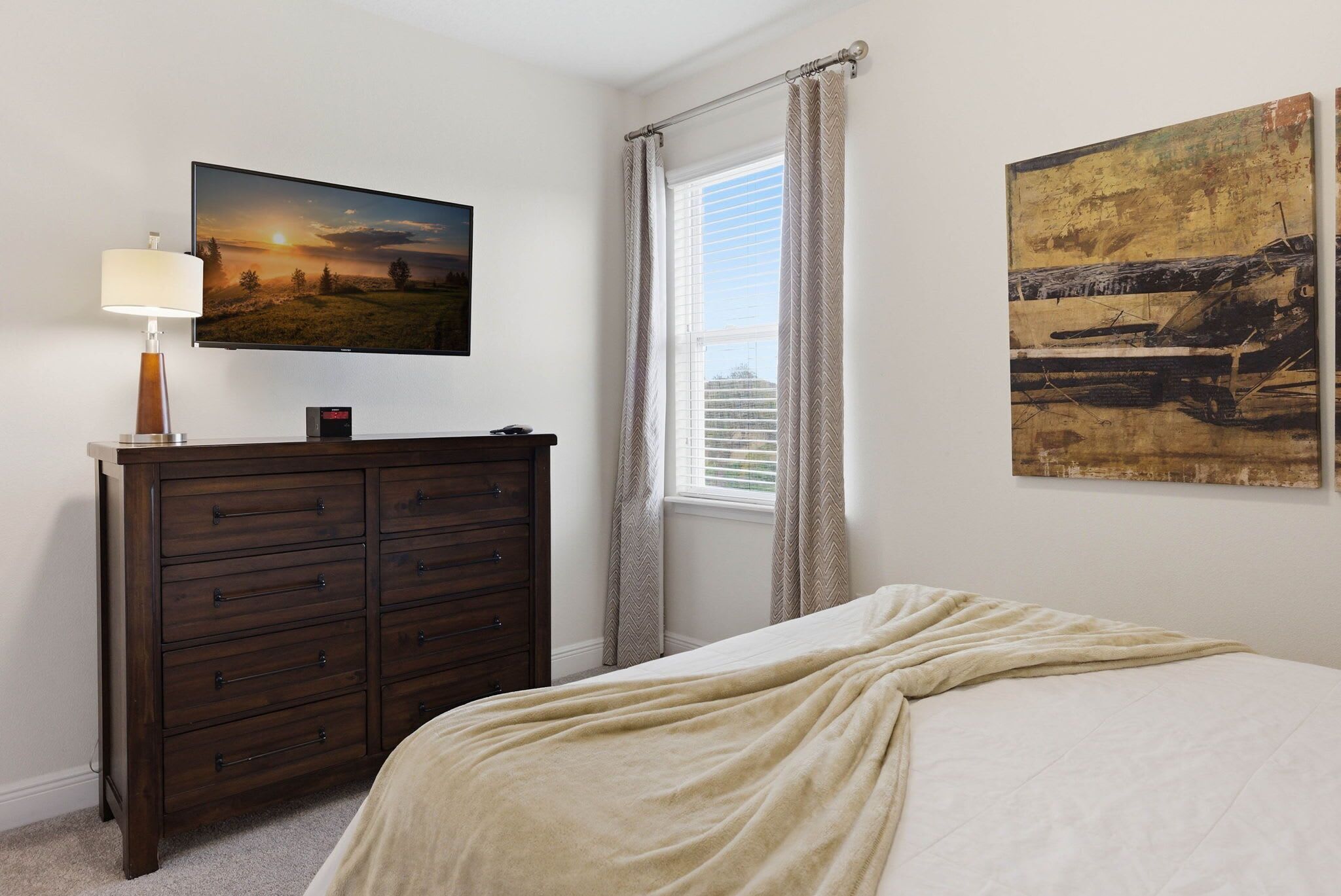 A bedroom with a bed, a dresser, a TV, a window with curtains, and two paintings.