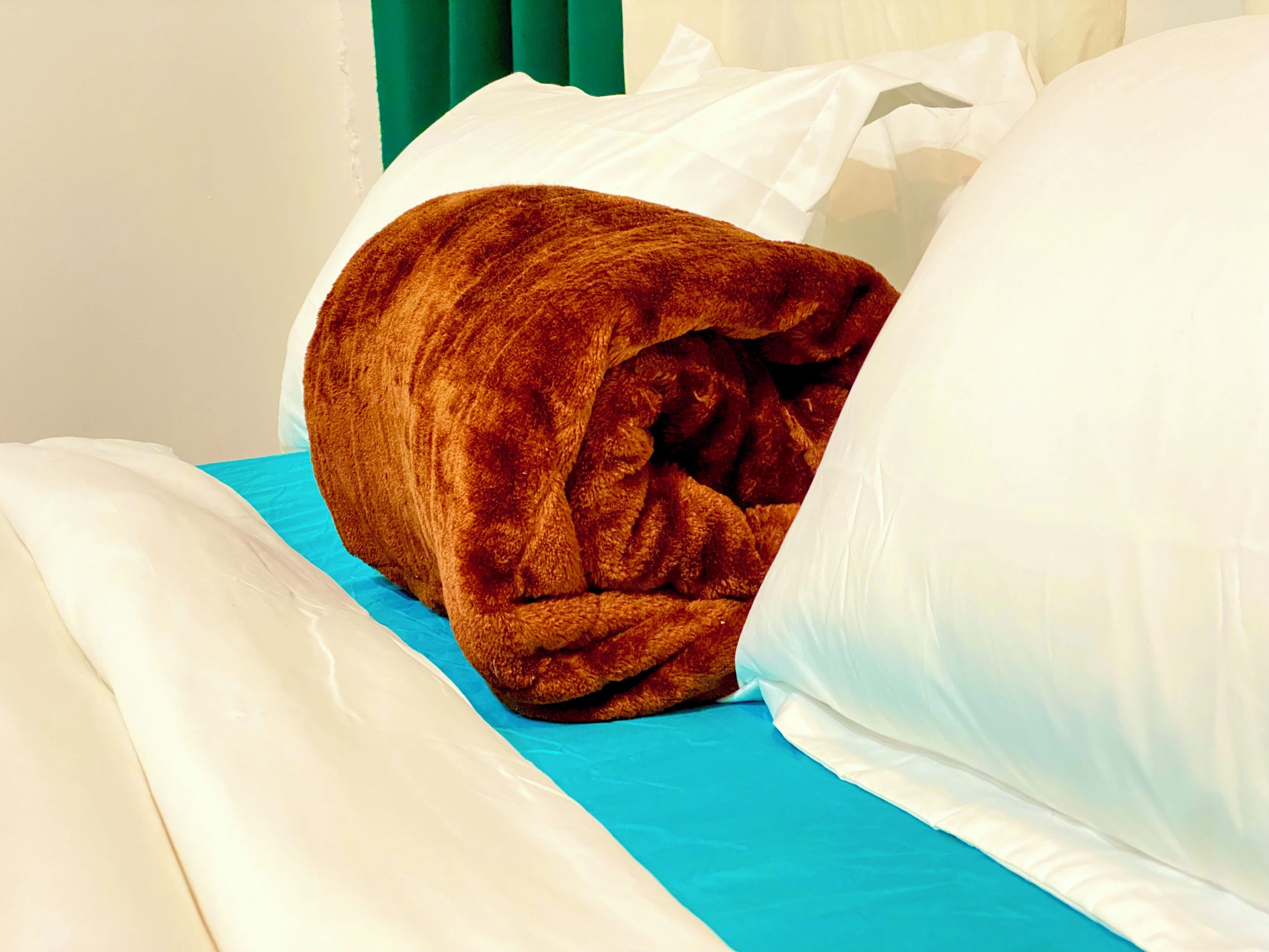Una cama con colchón azul, almohadas blancas y manta marrón.