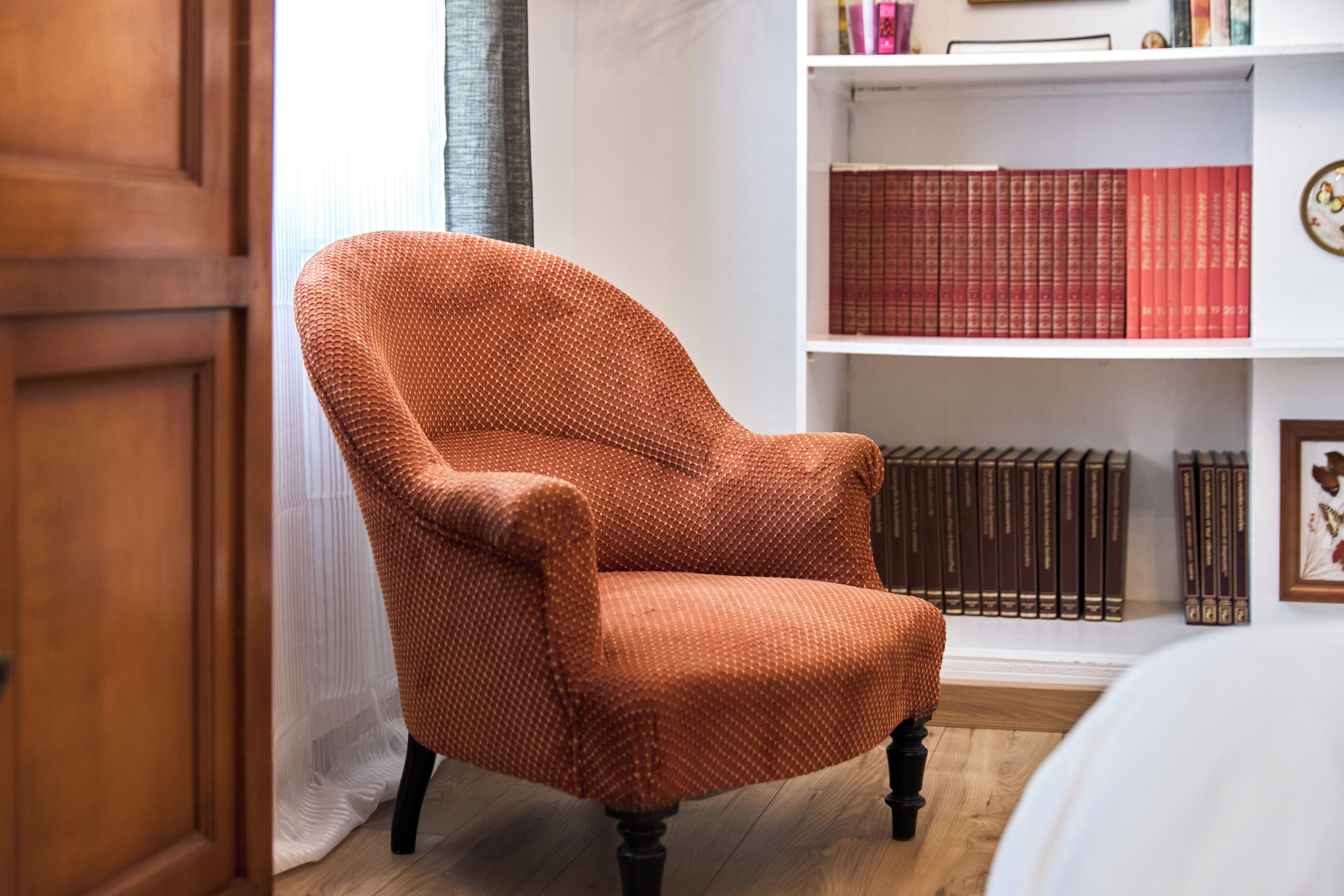 A room with a wooden door, a woven orange armchair, a white bookshelf with books, and a framed picture.
