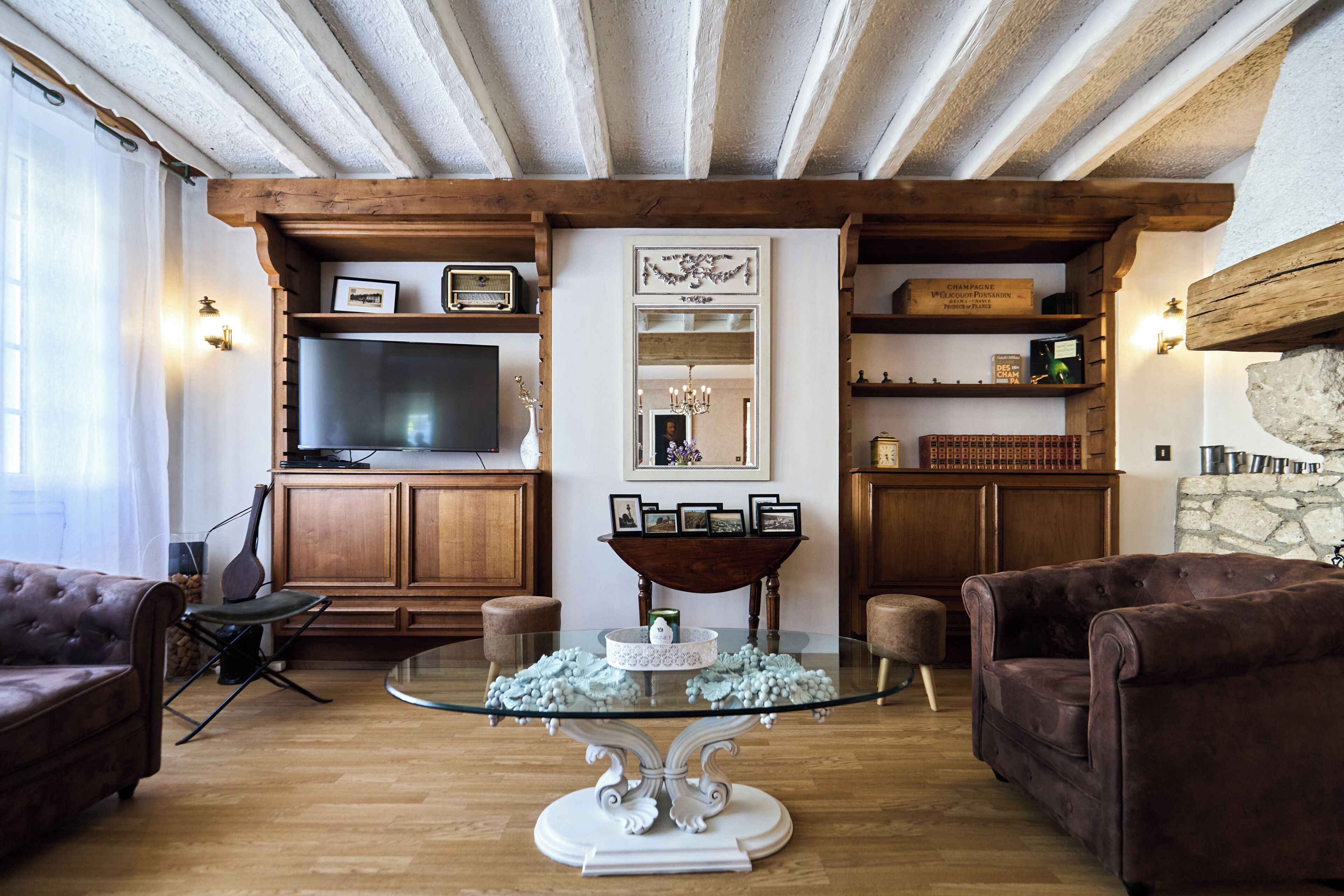 A living room with a glass coffee table, leather sofas, wooden beams, a television, and a stone wall.