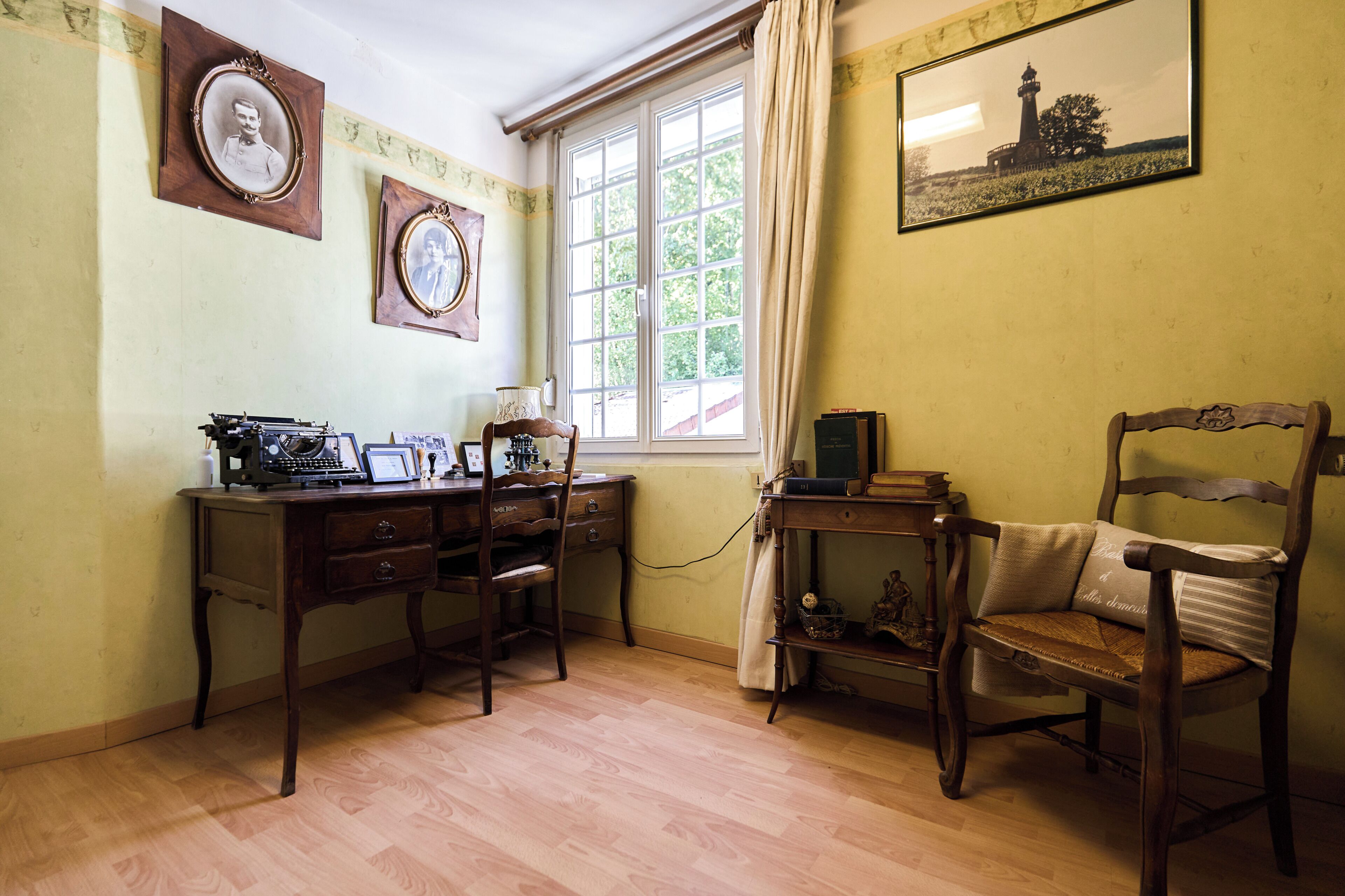 Una habitación con un escritorio de madera, una silla, una ventana con cortinas y cuadros en la pared.