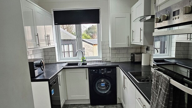 Una cocina moderna con armarios blancos, una lavadora negra, horno empotrado y una ventana con vista a una casa.