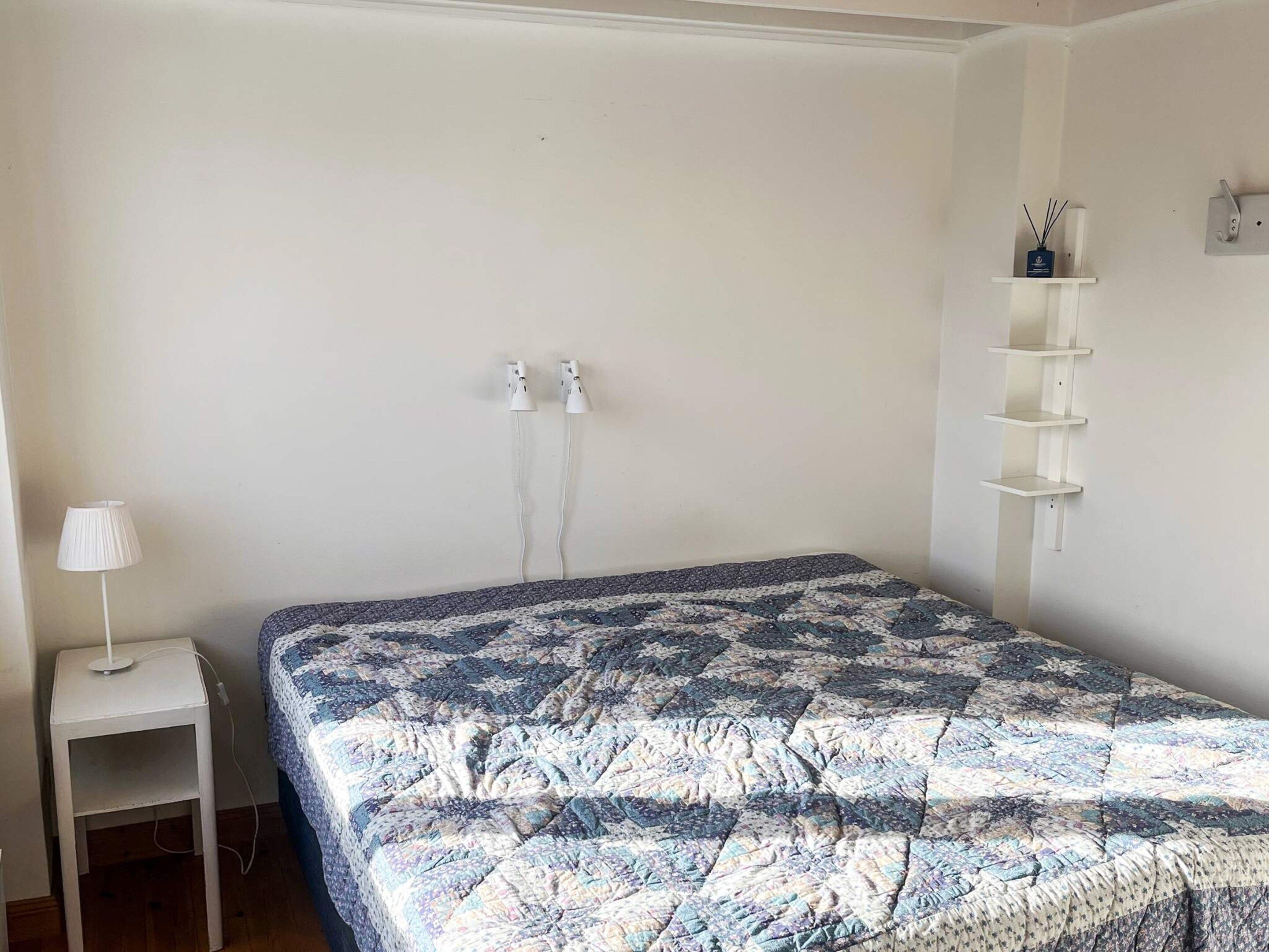 Un dormitorio con una cama, una mesita de noche con lámpara, un estante en la pared y dos enchufes.