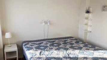 Un dormitorio con una cama, una mesita de noche con lámpara, un estante en la pared y dos enchufes.