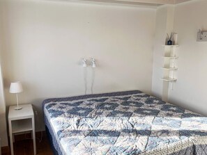 Ein Schlafzimmer mit einem Bett, einem Nachttisch mit Lampe, einem Wandregal und zwei Steckdosen.
