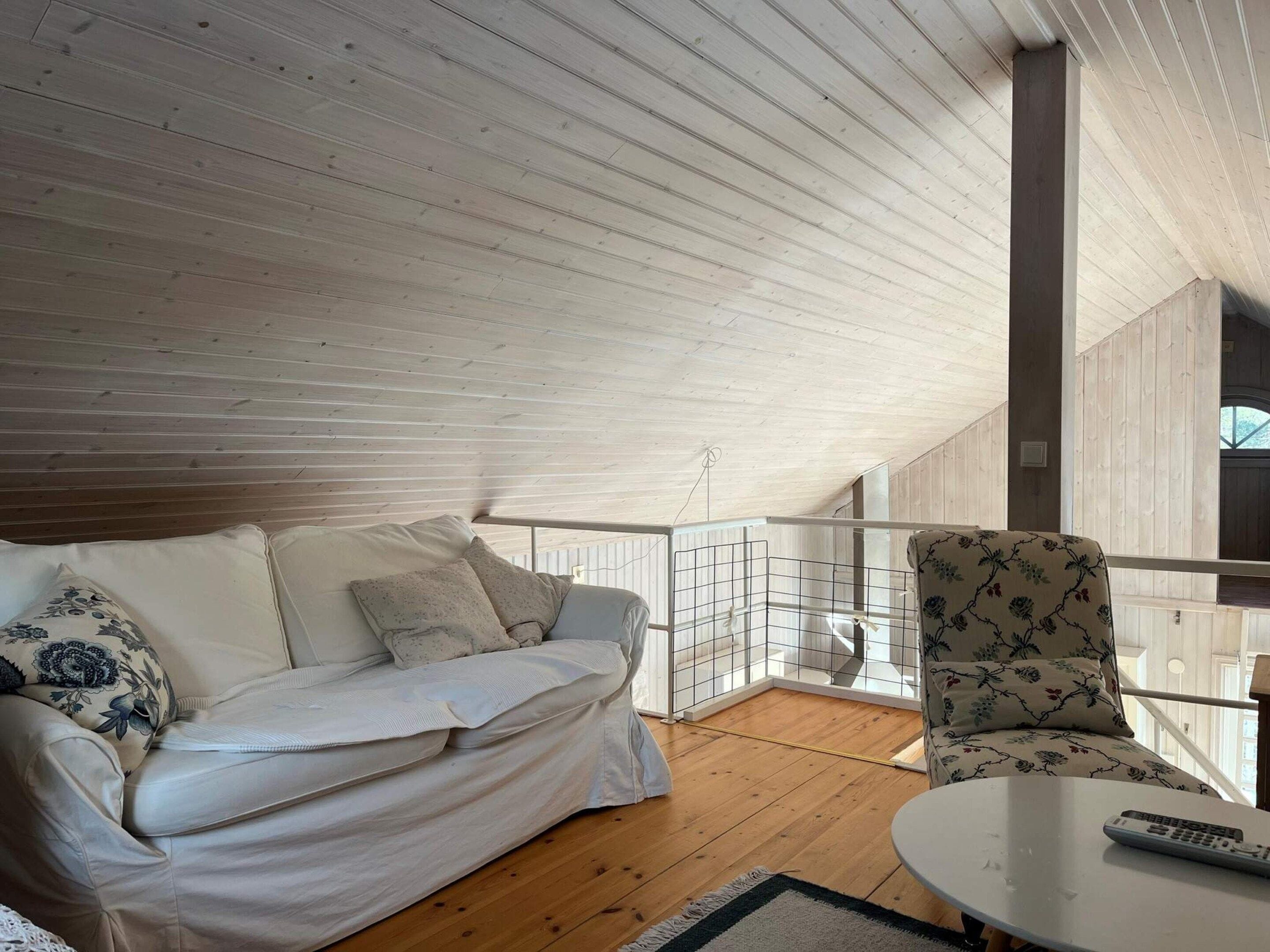 Quarto com teto de madeira, um sofá branco com almofadas estampadas, uma poltrona estampada e uma pequena mesa com um controle remoto.