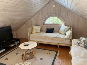 Ein gemütliches Dachzimmer mit Holzbalkendecke, einem Bett mit Strickdecke, einem kleinen runden Tisch, einem Fernseher und einem Fenster mit Blick auf Bäume.