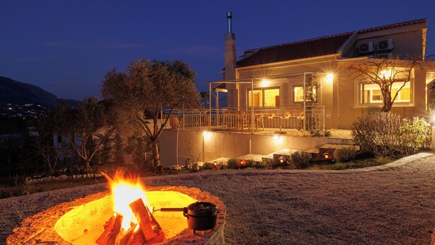 Ein Haus mit Feuerstelle im Vordergrund, einem überdachten Außenbereich mit Beleuchtung und einem Berg im Hintergrund.