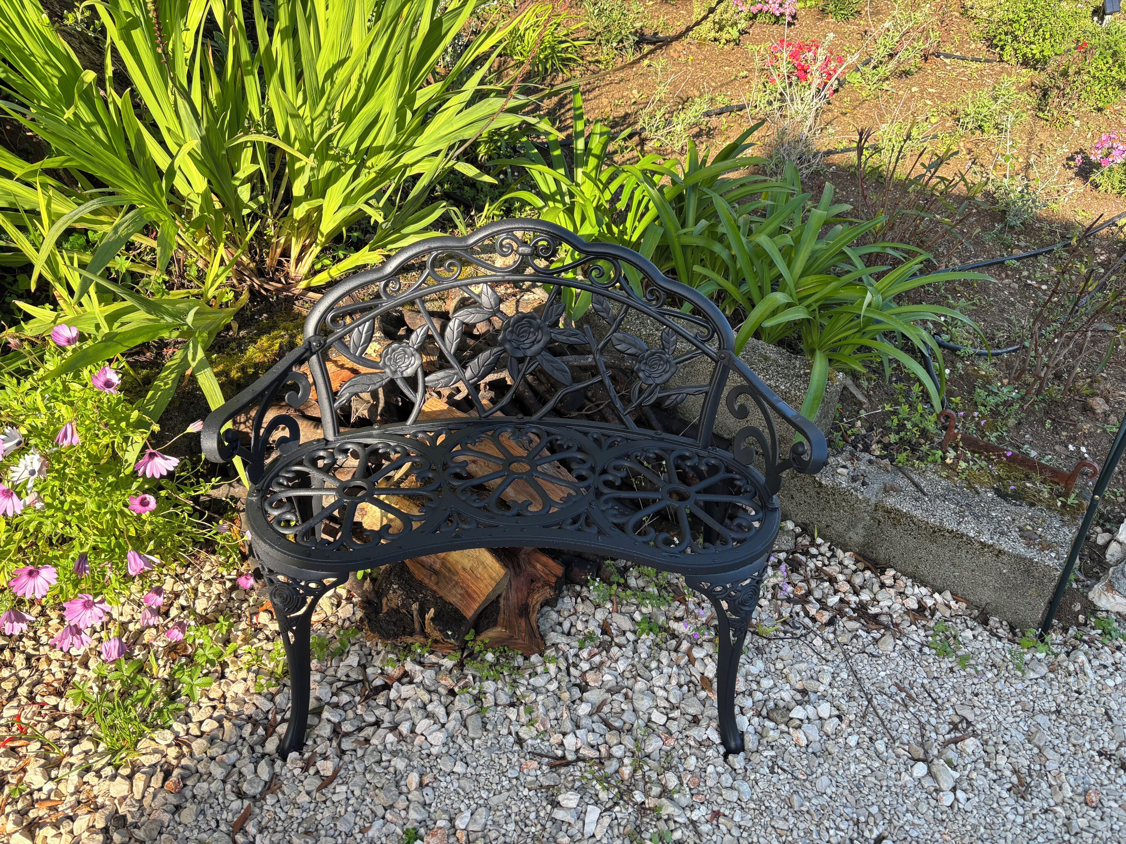 A black wrought iron bench with intricate designs, positioned on a gravel surface surrounded by greenery and flowers.