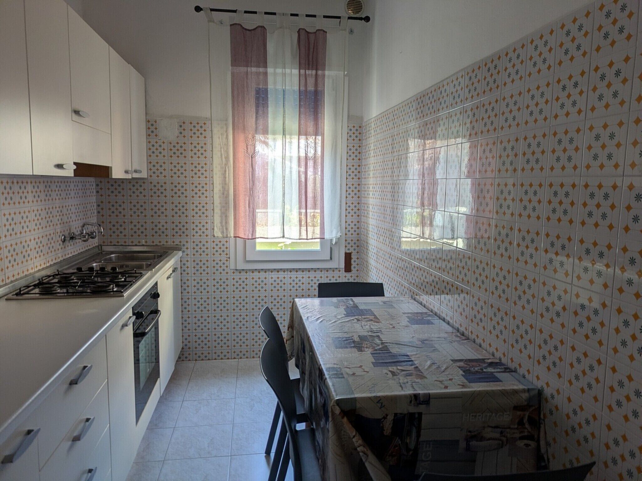 Una cocina con armarios blancos, una estufa a gas, una mesa de comedor con mantel de papel periódico y baldosas estampadas en las paredes.