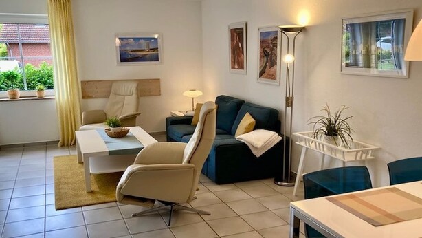 A modern living room with a sofa, armchair, coffee table, and a dining table. There are potted plants, artwork on the walls, and a window with curtains. - Ferienwohnung in Petersdorf auf Fehmarn (Petersdorf)