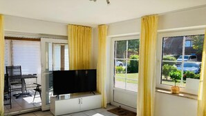 A room with a TV on a stand, a sliding door, a window with curtains, and a view of the outside. - Ferienwohnung in Petersdorf auf Fehmarn (Petersdorf)