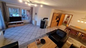 A living room with a TV, fireplace, dining area, and a view through large windows. - Ferienwohnung in Petersdorf auf Fehmarn (Petersdorf)