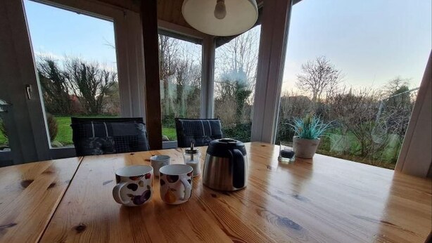 A wooden table with coffee cups and a kettle, large windows with a view of trees and a garden. - Ferienwohnung in Petersdorf auf Fehmarn (Petersdorf)
