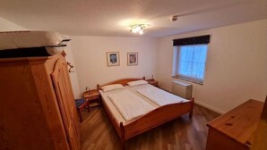 A bedroom with a wooden bed, bedside tables, a window with curtains, and a ceiling light fixture. - Ferienwohnung in Petersdorf auf Fehmarn (Petersdorf)