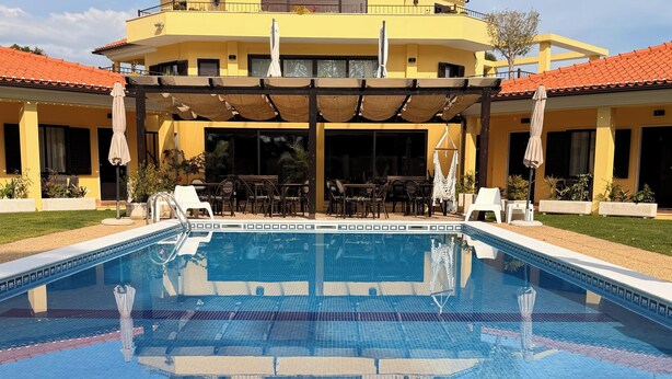 Una piscina con un borde de baldosas, mobiliario exterior y un edificio amarillo con techo rojo al fondo.