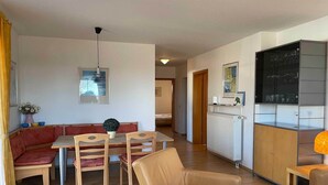 Un espace de vie compact comprenant un coin repas, un coin salon avec un canapé et des fauteuils, un coin cuisine avec un meuble de rangement et un radiateur.