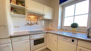 Une cuisine avec des meubles blancs, un lave-vaisselle Bosch, une cuisinière et une fenêtre agrémentée d’une plante.