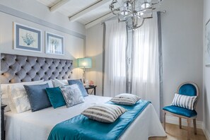 A bedroom with a tufted headboard, a bed with teal and white bedding, a wooden chair, and a chandelier. - Villa in Sardinia Near Porto Ottiolu Beach (Budoni)