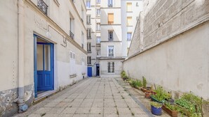 Une ruelle étroite avec une porte bleue, des plantes en pot et un immeuble doté de nombreuses fenêtres.