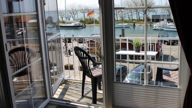A view from a room with a glass door to a balcony overlooking a marina with boats and a flag. - Ferienwohnung in Orth auf Fehmarn (Orth)