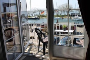 A view from a room with a glass door to a balcony overlooking a marina with boats and a flag. - Ferienwohnung in Orth auf Fehmarn (Orth)